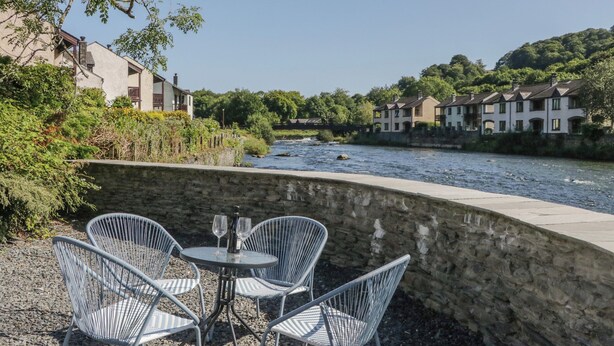 A riverside terrace with stone walls and metal chairs, overlooking a river and residential buildings. - Old Post Office Cottage (Ulverston)