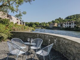 Terraza a la orilla del río con paredes de piedra y sillas de metal, con vista a un río y edificios residenciales.