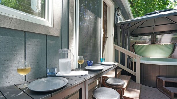 Een houten terras met een jacuzzi, een tafel met borden en glazen, en een barkruk.