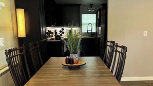 Une salle à manger avec une longue table en bois, des chaises noires, un grand vase central orné de plantes, et une cuisine en arrière-plan.