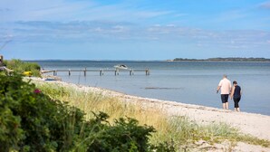 Ein Strand mit einer hölzernen Pier, die sich über ruhiges Wasser erstreckt, einem Boot, das am Ende liegt, und einer Person, die am Ufer entlanggeht.