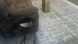 A living room with a brown sofa, a glass coffee table, a wooden side table, a refrigerator, a clock, a door, and a framed picture on the wall.