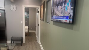 A hallway with a wooden floor, a clock, a television displaying a news program, and a bench.
