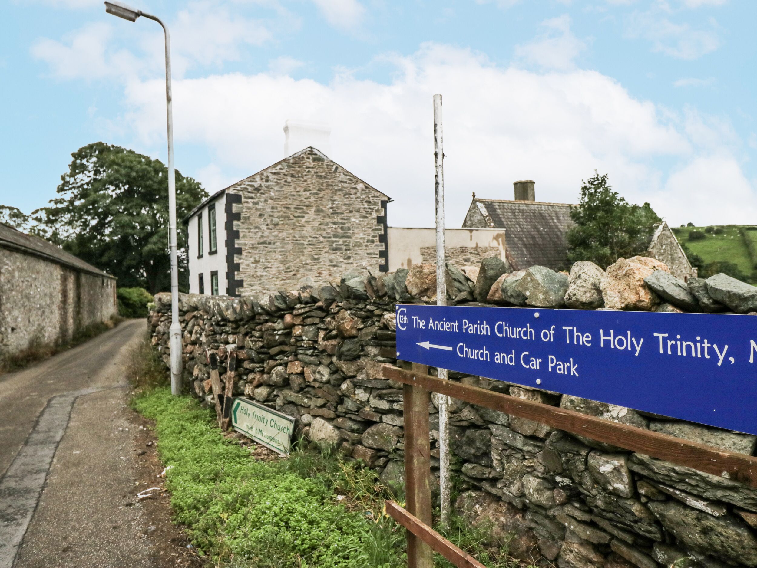 A rural road with stone walls and a sign for The Ancient Parish Church of The Holy Trinity, Church and Car Park.