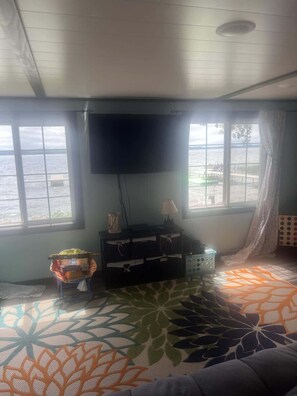 Ein Zimmer mit großem Fenster, einem flachbildfernseher an der Wand, einem gemusterten Teppich und einem kleinen Tisch mit einem Korb.