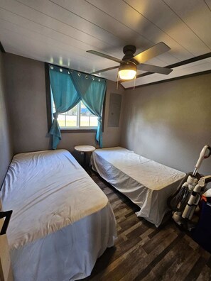 Une chambre avec deux lits, un ventilateur de plafond, une fenêtre avec des rideaux et une petite table.
