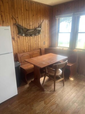 Une pièce aux murs lambrissés, avec une table et un banc en bois, un réfrigérateur blanc et une fenêtre.