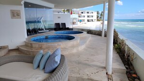 Un área de piscina con dos jacuzzis, una zona de estar con cojines y vistas a la playa y al océano.