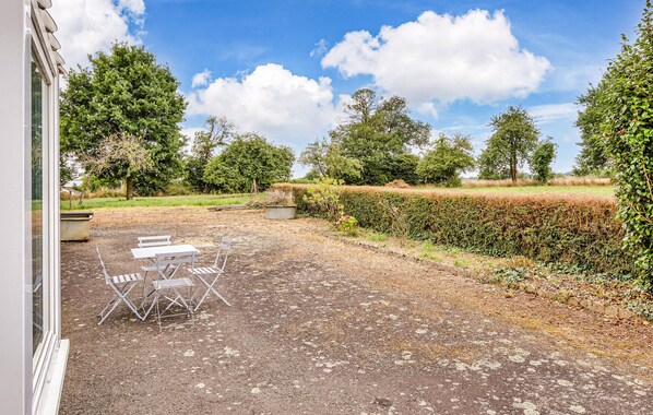 An outdoor patio area with a table and chairs, surrounded by a hedge and trees.