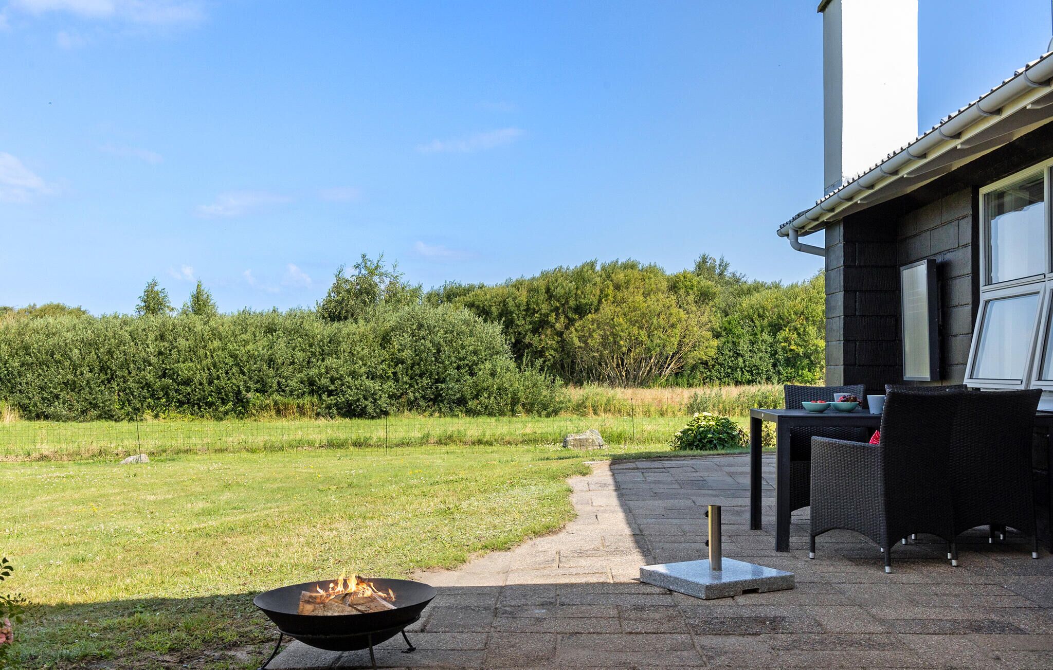 An outdoor patio with a fire pit, a dining area with wicker furniture, and a view of a grassy field and trees.
