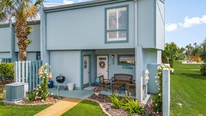 A two-story building with a light blue exterior, a front porch with a bench, and a small garden area with a palm tree and flowers.