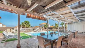 Un espace piscine extérieur avec une table et des chaises en verre, entouré d’une pergola et de palmiers.