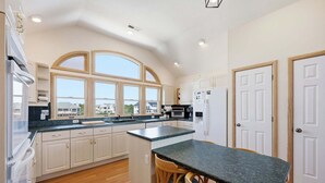 A kitchen with a central island, wooden cabinets, a built-in oven, and a view of houses through large windows. - 1397 - Sunshine Stars - Corolla Escape | Saltwater Pool & Hot Tub (Corolla)