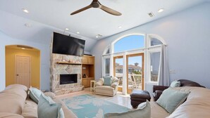 A living room with a stone fireplace, a flat-screen TV, a ceiling fan, and a large window with a view. - 1397 - Sunshine Stars - Corolla Escape | Saltwater Pool & Hot Tub (Corolla)