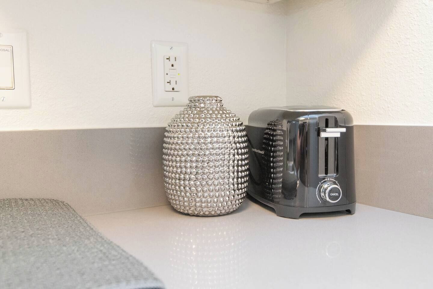 Una encimera blanca con un jarrón plateado decorativo y un tostador negro.