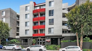 A multi-story building with a red balcony on the second floor, a visible entrance, and several parked cars.