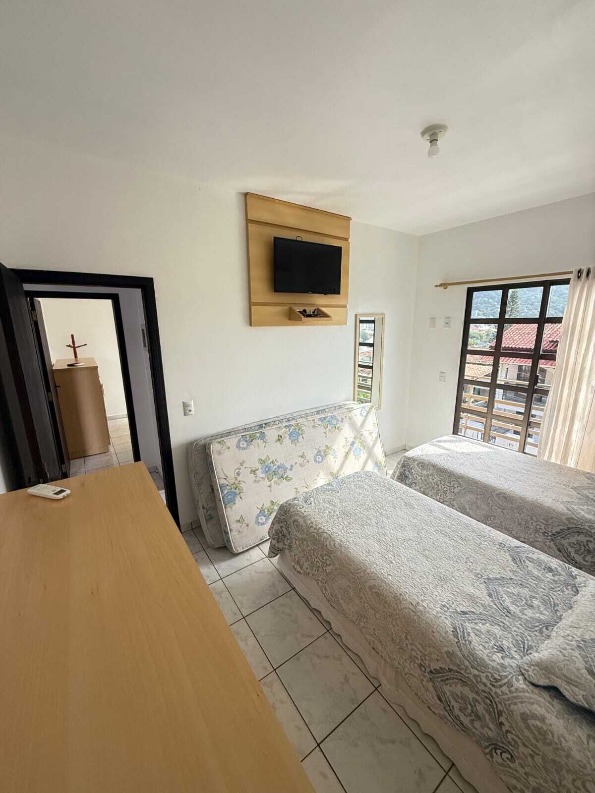 Ein Zimmer mit zwei Betten, einem an der Wand montierten Fernseher, einem Holztisch und einem Fenster mit Vorhängen.