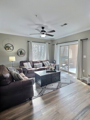 A living room with a brown leather sofa, a coffee table, and a ceiling fan. There is a window with curtains and a view of the outside.