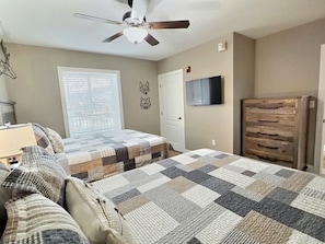 Een slaapkamer met een bed, een dressoir, een plafondventilator, een raam met jaloezieën en een televisie aan de muur.