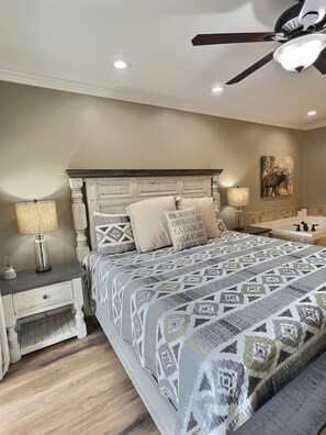 A bedroom with a large bed, two bedside tables, a ceiling fan, and a wall-mounted artwork.