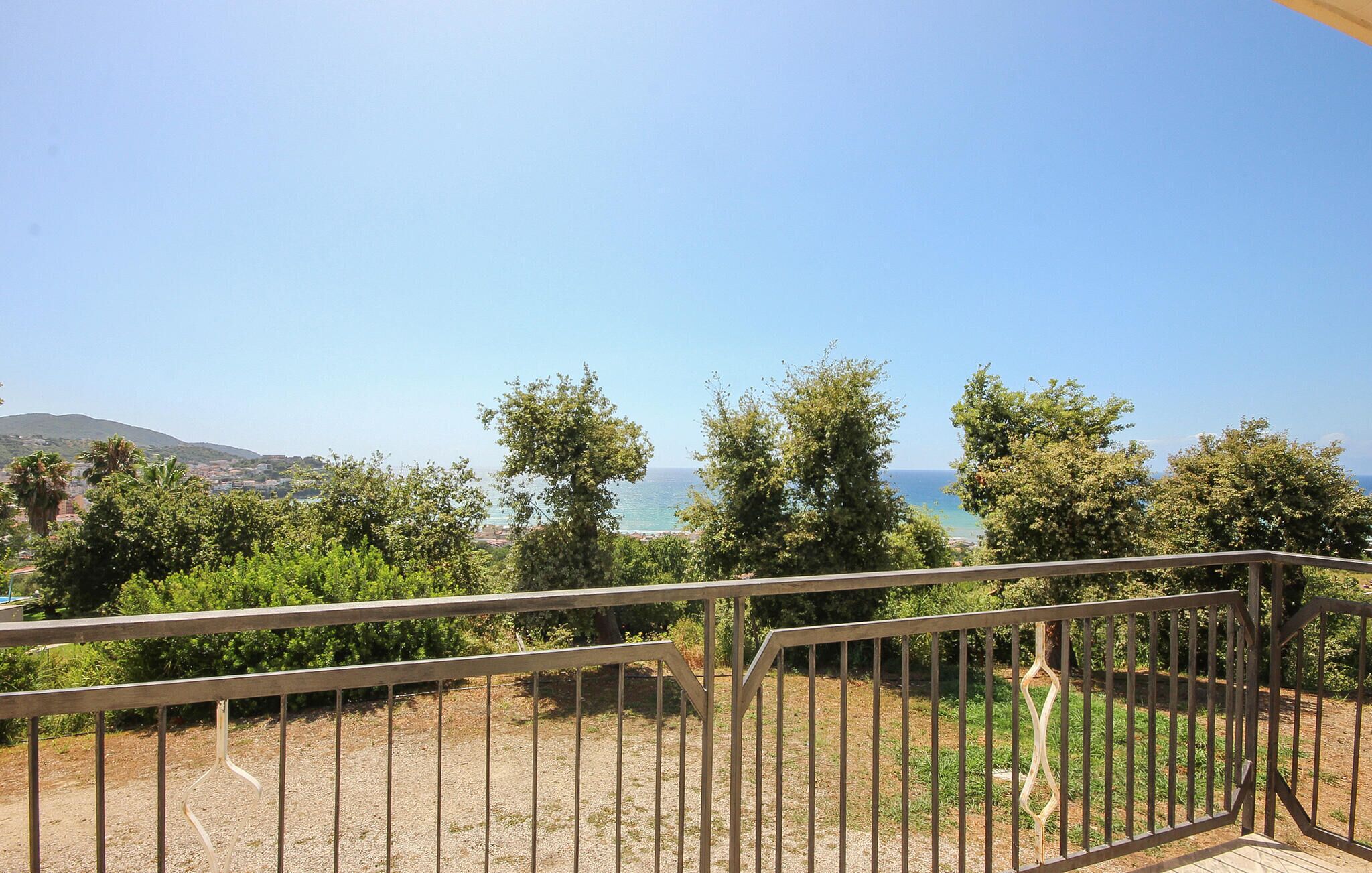 Un balcon doté d’une balustrade en métal, offrant une vue sur un paysage côtier avec des arbres et une perspective lointaine sur la mer.