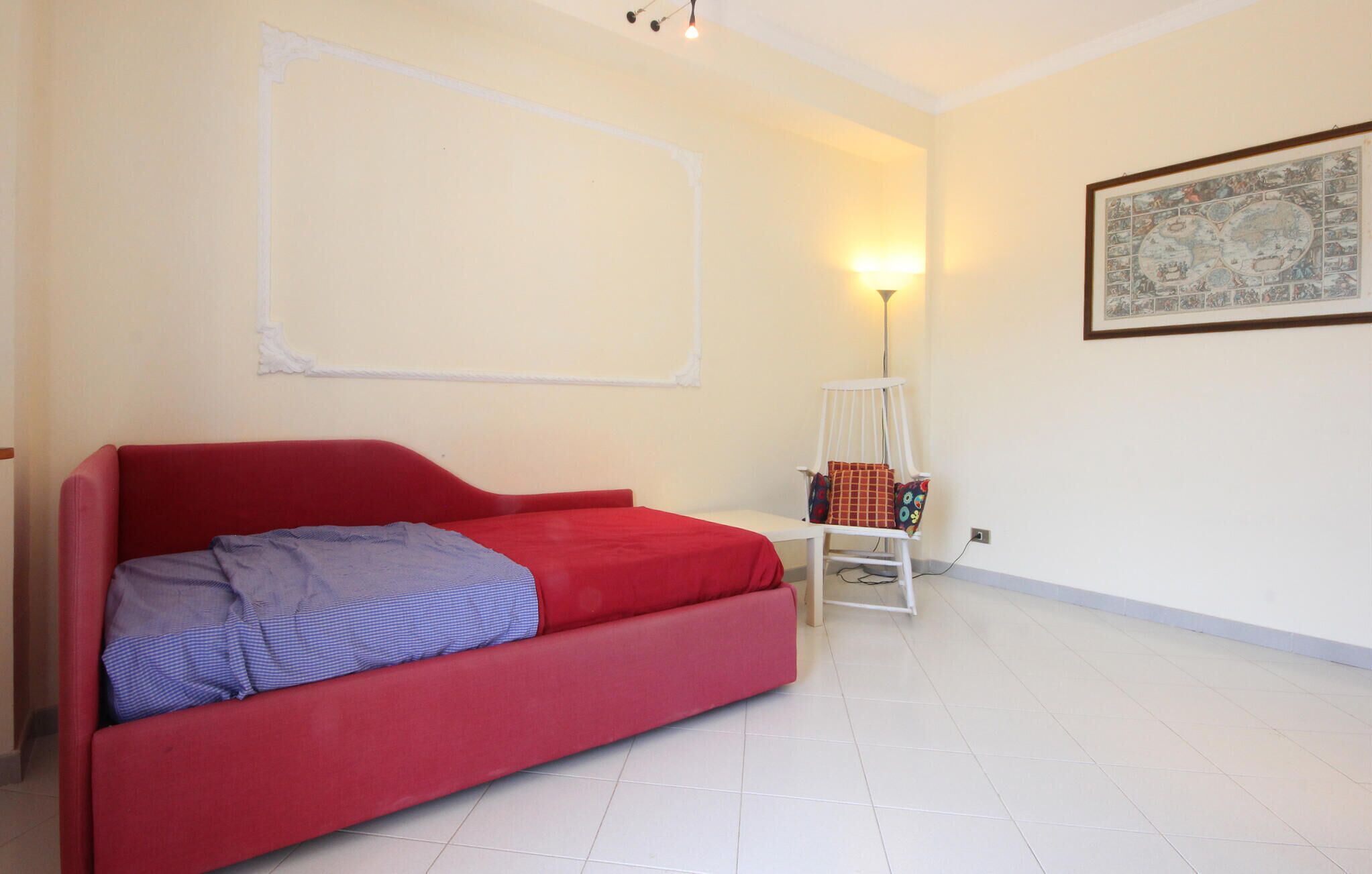 A room with a red sofa bed, a white chair, a small table, a wall-mounted lamp, and a framed map on the wall.