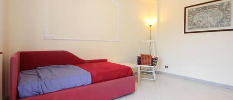 A room with a red sofa bed, a white chair, a small table, a wall-mounted lamp, and a framed map on the wall.