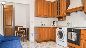 A compact kitchen with wooden cabinets, a washing machine, and a dining area with a blue sofa and wooden chairs. - 1 bedroom awesome apartment in Capaccio Paestum (Capaccio Paestum)