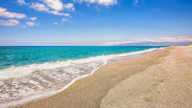 A beach with a pebbly shore and clear turquoise waters under a blue sky with scattered clouds.