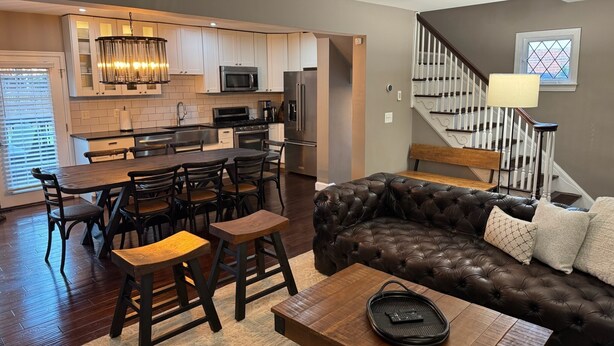 A modern kitchen with white cabinets, a dining area with a wooden table and chairs, and a leather sofa in a living room with a staircase and window. - Charming 2-bedroom, 2-bathroom house in peaceful Mount Rainier. (Mount Rainier)