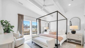A modern bedroom with a four-poster bed, a white sofa, a bedside table, and a large window with curtains.