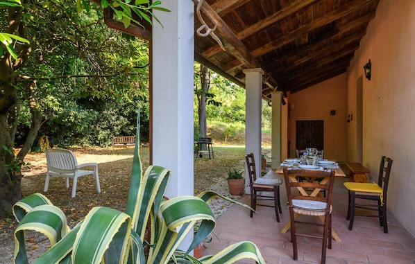 An outdoor patio with a wooden table set for dining, surrounded by chairs, and a covered area with a view of a garden.