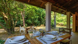 Un área de comedor al aire libre con una mesa de madera dispuesta para cuatro personas, rodeada de exuberante follaje y una estructura cubierta.