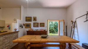 Un comedor amplio con una gran mesa de madera, una cocina con pared de ladrillo y vistas a la vegetación a través de una puerta abierta.