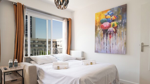 Une chambre d’hôtel avec un grand lit, une table de chevet avec une télécommande, une commode, une fenêtre avec des rideaux et un tableau représentant des parapluies colorés.
