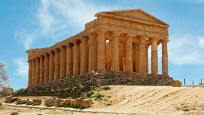 An ancient Greek temple with a series of columns and a pediment, situated on a hillside with a clear sky above.