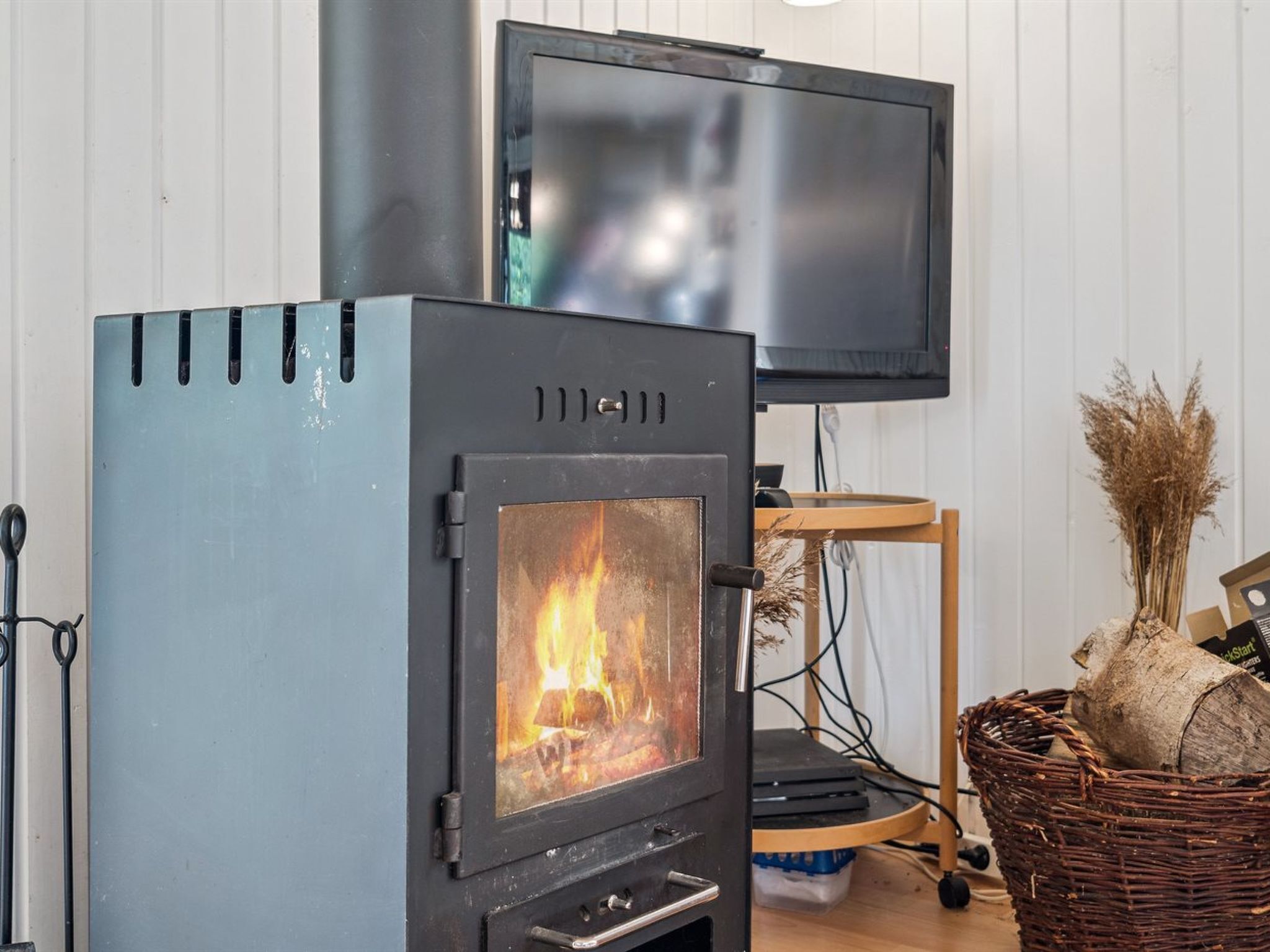 A wood-burning stove with a visible fire, a television mounted on the wall, and a wicker basket with logs on a table.