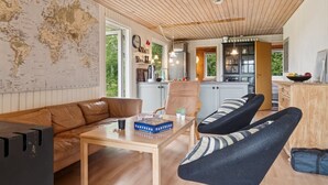 A spacious living room with a wooden ceiling, a world map on the wall, a leather sofa, a coffee table, and a dining area with a kitchen in the background.