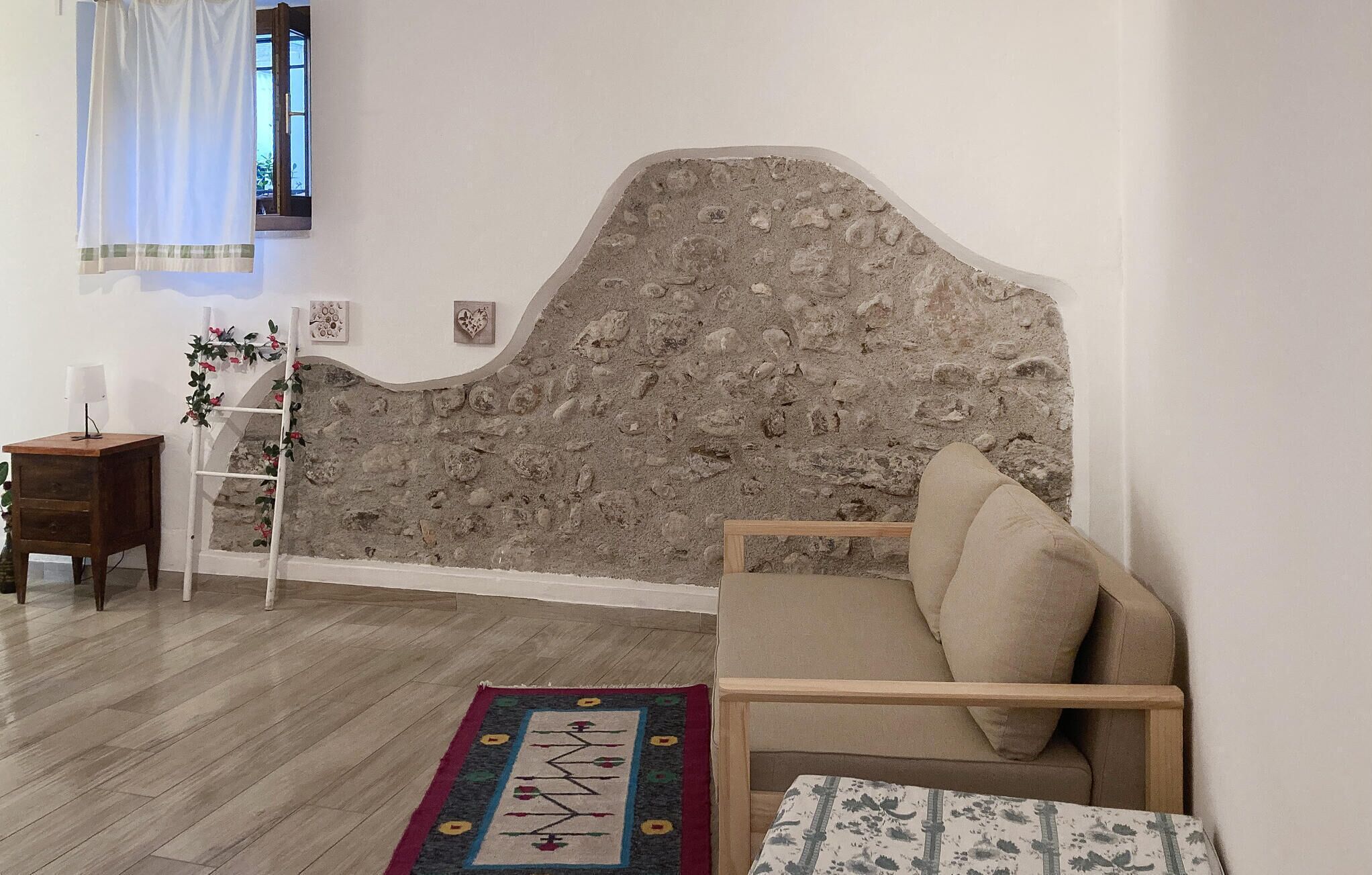 A room with a stone wall headboard, a wooden sofa, a small table with a lamp, a wooden ladder, and a patterned rug.