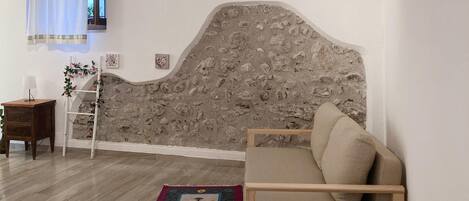 A room with a stone wall headboard, a wooden sofa, a small table with a lamp, a wooden ladder, and a patterned rug.