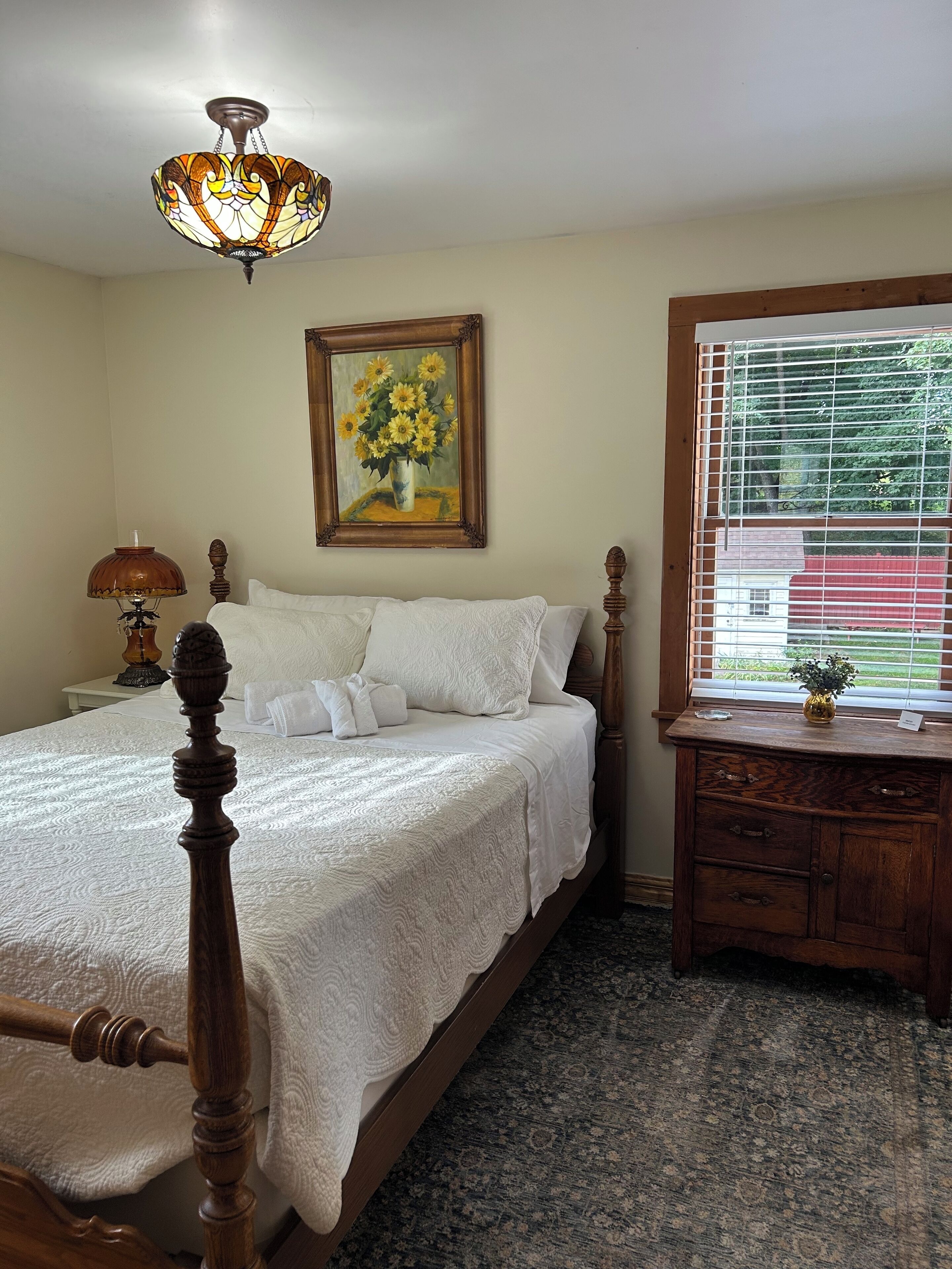 A bedroom with a four-poster bed, a nightstand with a lamp, a dresser, a painting of sunflowers, and a window with blinds.