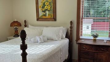 A bedroom with a four-poster bed, a nightstand with a lamp, a dresser, a painting of sunflowers, and a window with blinds.