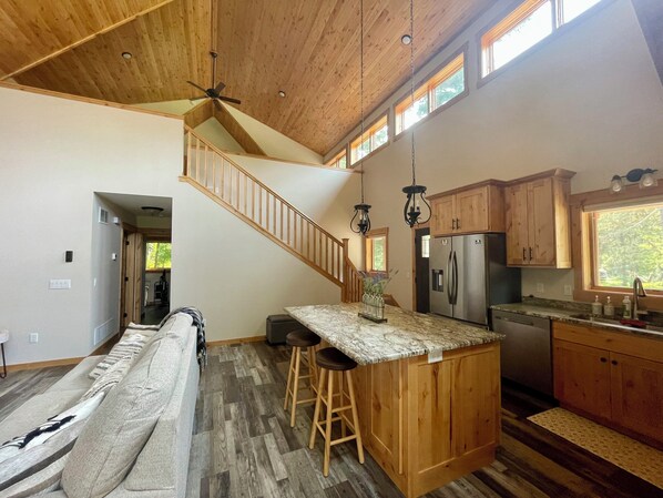 Una cocina amplia con armarios de madera, una isla central con taburetes, un refrigerador de acero inoxidable y una escalera de madera.