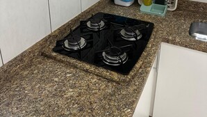 A kitchen with a gas stove, a trash bin, and a window with a fruit basket curtain.