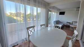 A room with a round table and chairs, a sofa, a TV mounted on the wall, and large windows with sheer curtains. - CAP D'AGDE - Plage du Môle / Réf 405 (CAP D'AGDE)