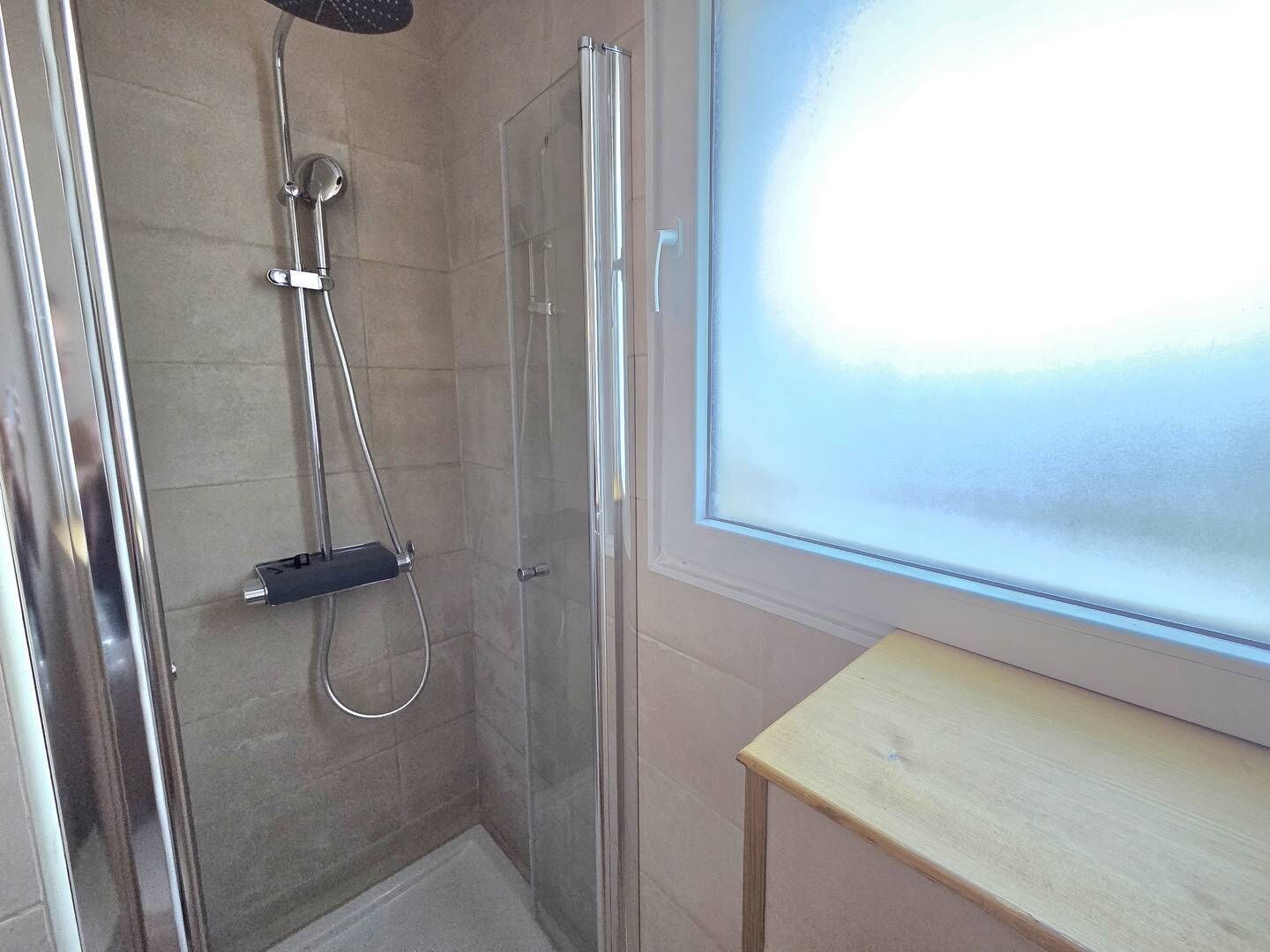 A shower with a glass door, a wooden bench, and a frosted window.