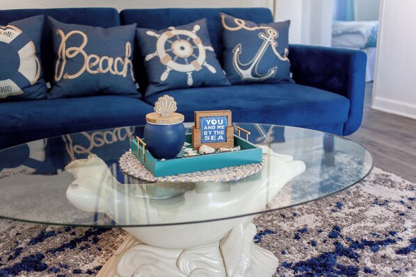 A glass-top coffee table with a decorative centerpiece featuring a small ship model and a sign that reads "YOU AND ME BY THE SEA."