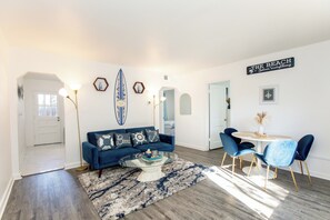 Una sala de estar con un sofá azul, una mesa de centro y un comedor con sillas y una mesa. Se observa una decoración con una tabla de surf y un letrero que dice "The Beach".