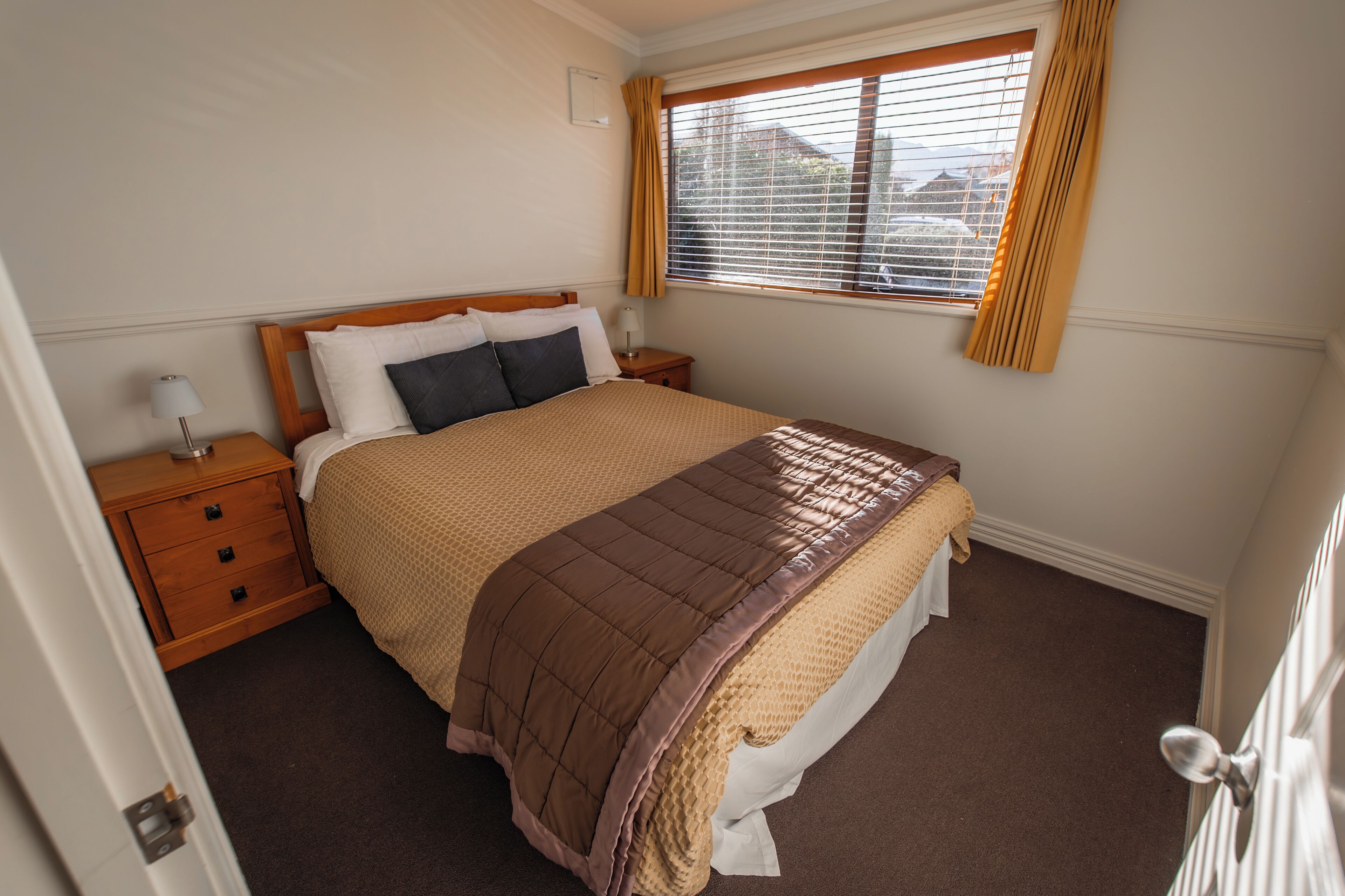 A neatly made bed with a brown quilt, a wooden nightstand with a lamp, and a window with blinds.