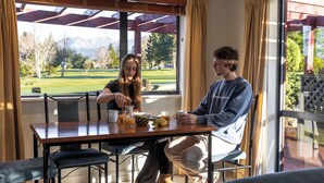 A dining area with a wooden table, chairs, and a view of a golf course through large windows. - Alpine Springs Motel | Hot Tub | Unit 2 - HS51212 (Hanmer Springs)